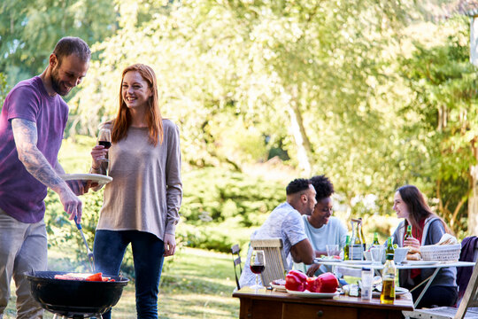 Young man preparing food in barbecue grill while his friends have a drink in background