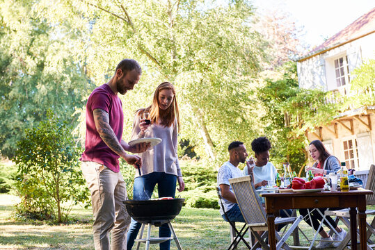 Young man preparing food in barbecue by his friends sitting at table