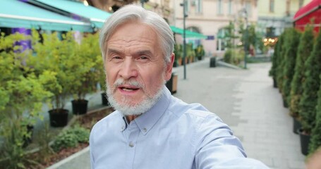 Selfie camera view of the senior bearded man talking via video call with his relatives while spending time at the street and telling something emotionally