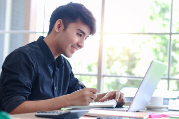 Asian young handsome businessman in black shirt working on office desk, taking notes and using laptop computer, writing information data and new idea on paper