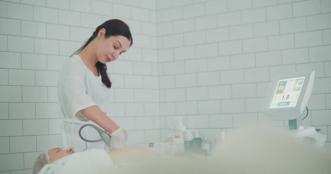 A close-up female cosmetologist is doing a SMAS lifting procedure on the female client by using the Ultraformer apparatus.