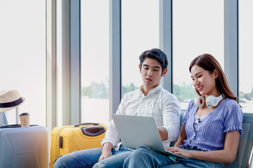 Asian young traveler lover couple siting on terminal chair seat with luggage suitcase, using laptop computer together, talking having and having fun times, happy friend traveling on vacation.