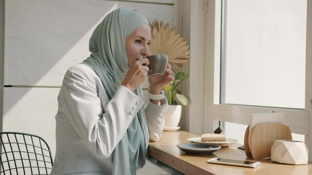 Slow motion of cheerful young Arab woman wearing hijab drinking tea enjoying leisure time in beautiful cafe. Muslim people and happiness concept.