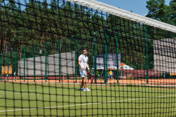 Young sportsman holding tennis racket near blurred net on court