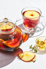 Fruit tea with apples and thyme and honey in glass teapot and cup on white background with hard shadows