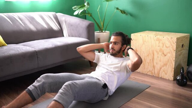 Latin man doing crunches with a twist while lying on an exercise mat in his living room