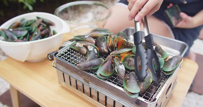 Woman roasting fresh juicy Green clam Shell mussels with tongs grilling on a hot stove, cooking seafood party outdoor