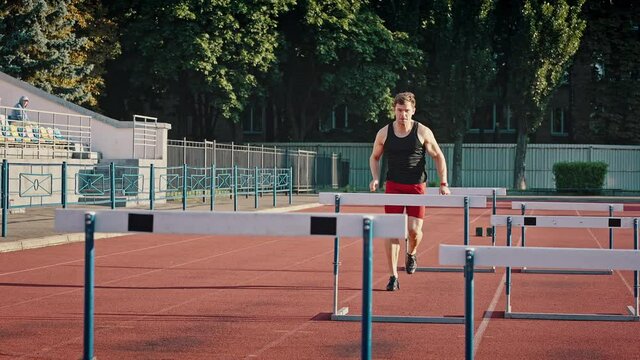 Active guy athlete in sports shorts and a T-shirt runs a distance jumping over obstacles. Competitions at the city stadium