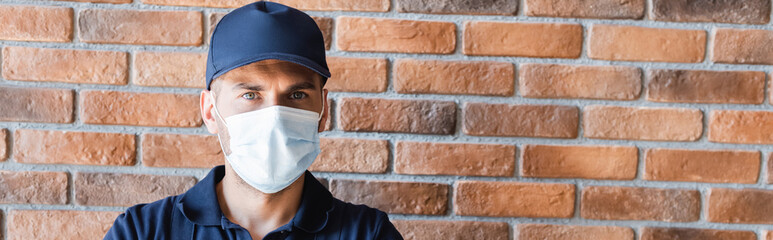 workman in cap and medical mask looking at camera near brick wall, banner