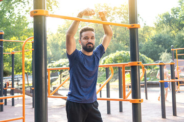 Obraz premium Open air gym. Street training on the municipal sports equipment. Athletic bearded sportsman posing near the horizontal bar in the city park.