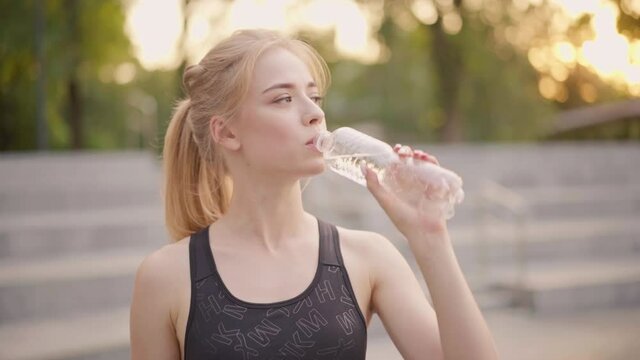 Sport and Fitness. Young Caucasian Woman Dressed Sportswear Standing Outdoor Drink Water Plastic Bottle Summer Park Sunlight Background Resting After Training Jogging Running Exercises loosens hair