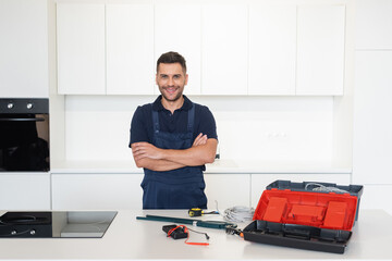 smiling repairman standing with crossed arms near tools and wires in kitchen