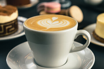 Café  con arte latte en un primer plano acompañado de distintos tipos de tortas con foco diferenciado.