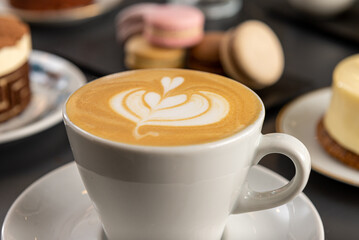 Café  con arte latte en un primer plano acompañado de distintos tipos de tortas con foco diferenciado.