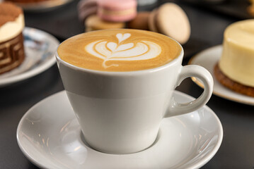 Café  con arte latte en un primer plano acompañado de distintos tipos de tortas con foco diferenciado.