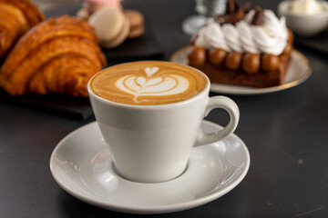 Café  con arte latte en un primer plano acompañado de distintos tipos de tortas con foco diferenciado.