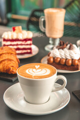 Café  con arte latte en un primer plano acompañado de distintos tipos de tortas con foco diferenciado.