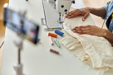 African-American dressmaker sews white fabric with new machine at table in studio