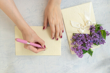 Obraz na płótnie Canvas congratulations concept. woman's hands write on blank card or letterhead near purple flowers in beautiful envelope with bow. Beautiful lilac flowers. Flat lay with spring bloom. top view