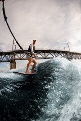 adult man holds rope and rides wave on wakeboarding board against the background of bridge