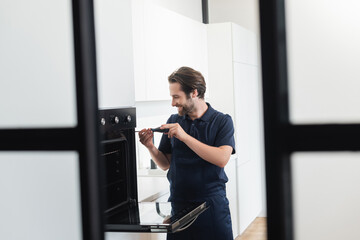 smiling handyman repairing inbuilt electric oven with screwdriver on blurred foreground