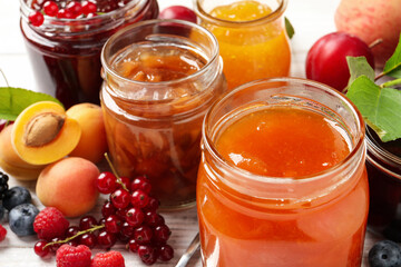Jars with different jams and fresh fruits on white wooden table, closeup