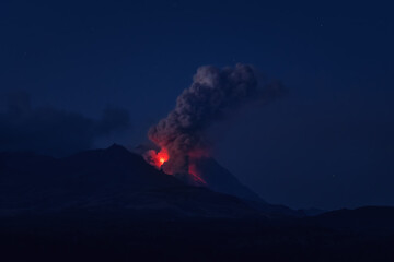The eruption of the Shiveluch volcano on the Kamchatka Peninsula, Russia. Summer night landscape with ash-gas ejection of a volcano, red flowing lava, starry sky.