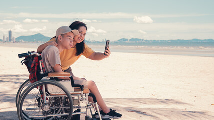 Happy mother and disabled teenage boy on wheelchair use social media, Sea beach...