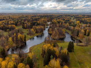 Autumnal forest with golden crowns of trees. Colorful public park with lake. Foliage of trees change color from green to yellow and orange. Promenade. Cloudy sky. Change of season concept. Aerial view