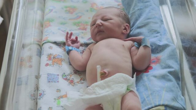 A newborn baby with maternity hospital bracelets on his arm is lying restlessly in the crib