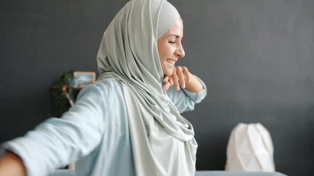 Tilt-down slow motion portrait of happy young muslimah wearing hijab dancing having fun indoors in apartment. Lifestyle and modern youth concept.