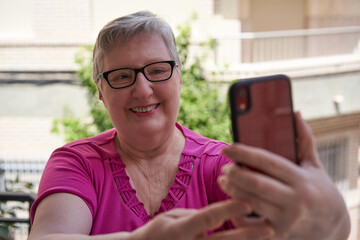 Older woman in pink t-shirt on her terrace at home. She is holding a red smartphone and taking a selfie.