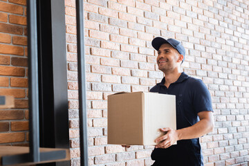 cheerful delivery man holding cardboard package near brick wall