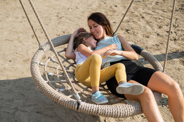 Mom and daughter swing on a round swing. Caucasian woman and little girl have fun on the playground.