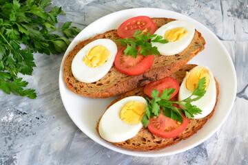 sandwiches made of whole grain bread with boiled eggs, tomato slices and parsley on white plate on concrete background