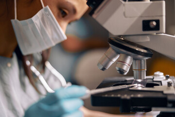 Asian technician with mask drips liquid sample onto slide working with microscope in clinic