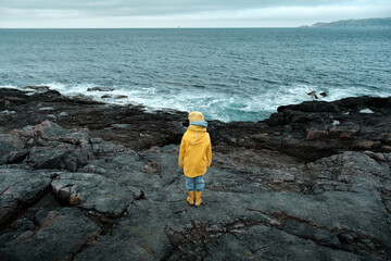 A child in a yellow raincoat looks at the ocean on a cold cloudy day