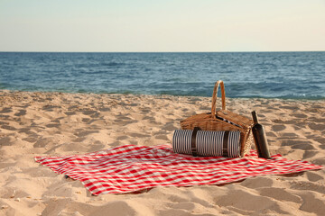Blanket with picnic basket and bottle of wine on sandy beach near sea, space for text
