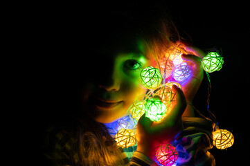portrait of a little seven year old kid girl holding a garland in her hands near her face, glowing with colorful multicolored neon lights at night time at home. Christmas Eve holiday celebration