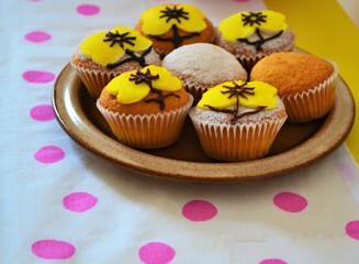 Plate of muffins decorated with powdered sugar and yellow icing in the shape of yellow flowers on a white tablecloth with pink polka dots on a yellow table
