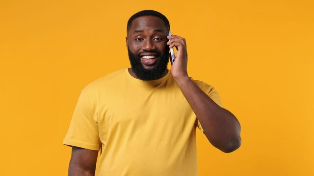 Happy calm cheerful young bearded african american man 20s wears orange t-shirt hold use talk on mobile cell phone conducting pleasant conversation isolated on plain yellow background studio portrait