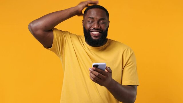 Fun young african american man 20s wear orange t-shirt hold use mobile cell phone say wow yes just found out great big win news doing winner gesture isolated on plain yellow background studio portrait