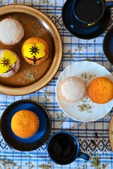 Blue coffee set with yellow flowers surrounded by muffins decorated with powdered sugar and icing in the shape of a yellow flower that matches those of the coffee set on a white and blue squares