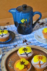 Blue coffee set with yellow flowers surrounded by muffins decorated with powdered sugar and icing in the shape of a yellow flower that matches those of the coffee set on a white and blue squares
