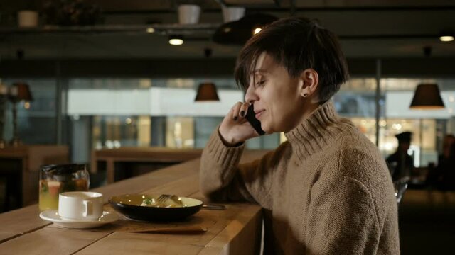 a cute girl in a beige warm sweater talking on the phone and laughing merrily sitting in a restaurant. High quality FullHD footage