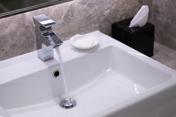 Closeup faucet with flowing water, white sink, and soap. Lavatory grey marble restroom interior concept.