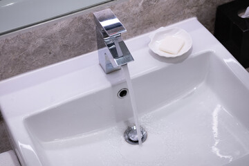 Closeup faucet with flowing water, white sink, and soap. Lavatory grey marble restroom interior concept.