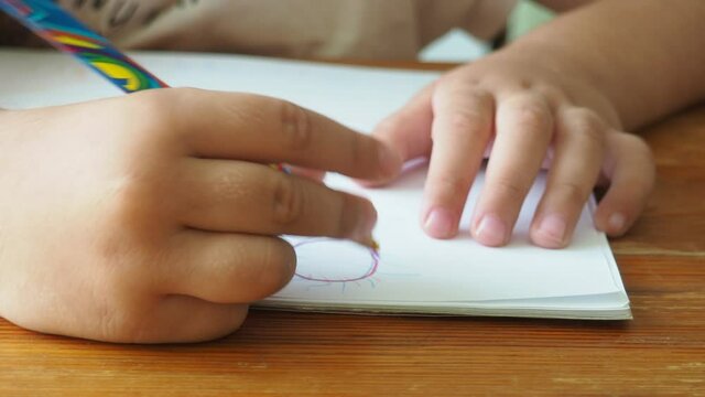 Cute little preschooler child drawing at home. close-up of a little girl's hand. holds a pencil in his hands. child draws a house, clouds sun. multicolored rainbow pencil