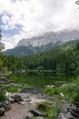 Obraz premium Ein kleiner grüner Teich, umgeben von einem Wald und Steinen. Im Hintergrund ist ein von Wolken umgebener Berg zu sehen. Die Bäume spiegeln sich im Teich.