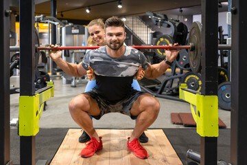 Attractive young fit woman assisting to muscular sportsman during strength training at gym. Man doing reverse push up over exercise bench with weight plate and sportswoman helping him.